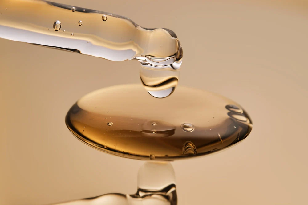 Close-up of a glass dropper dispensing a golden peptide serum droplet onto a reflective surface